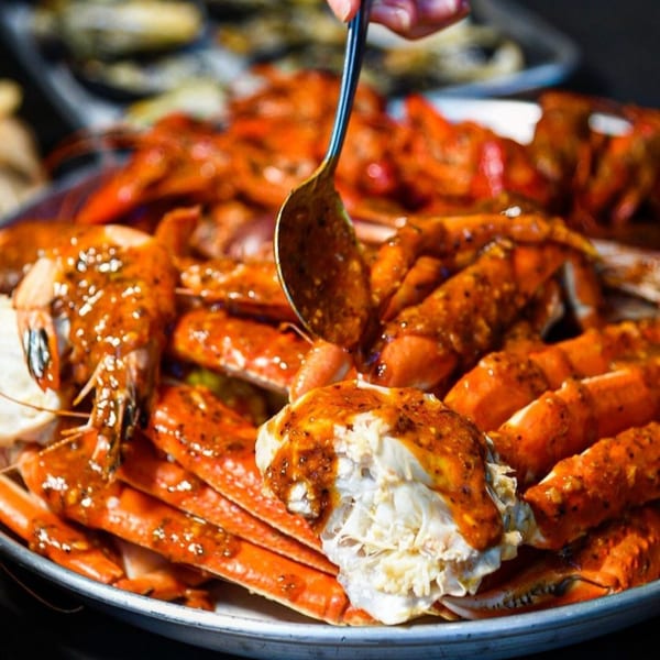 A spoon drizzles sauce over a platter of cooked crab legs and shrimp at The Crab Station.